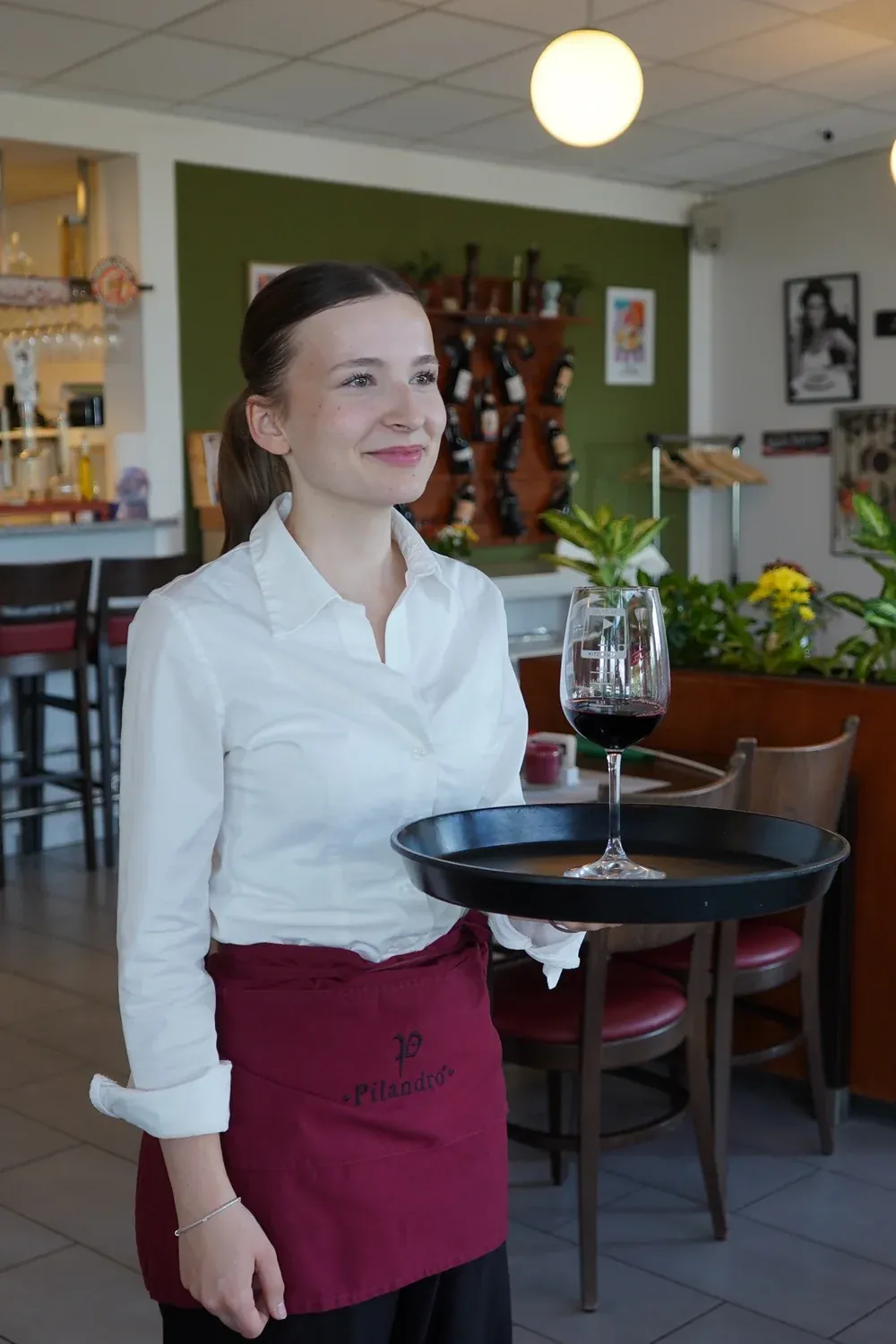 Eine Servicekraft im weißen Hemd trägt ein Glas Rotwein auf einem Tablett durch den Gastraum