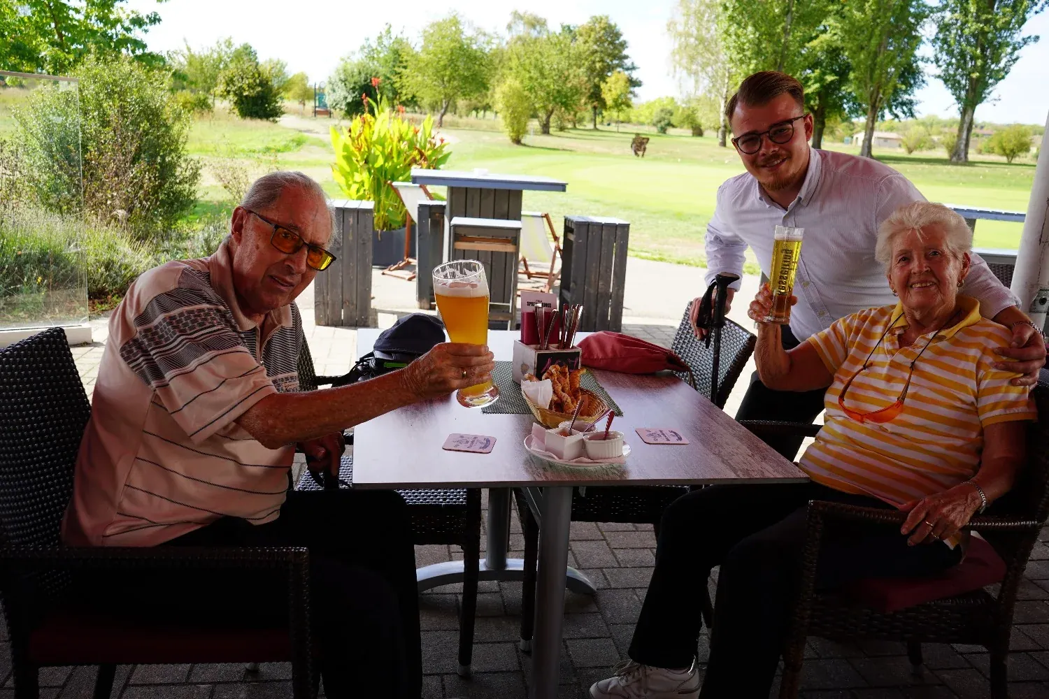 Ein älteres Paar stößt mit dem Kellner auf der Terrasse an, Weißbier und Pils in der Hand, dahinter der Platz