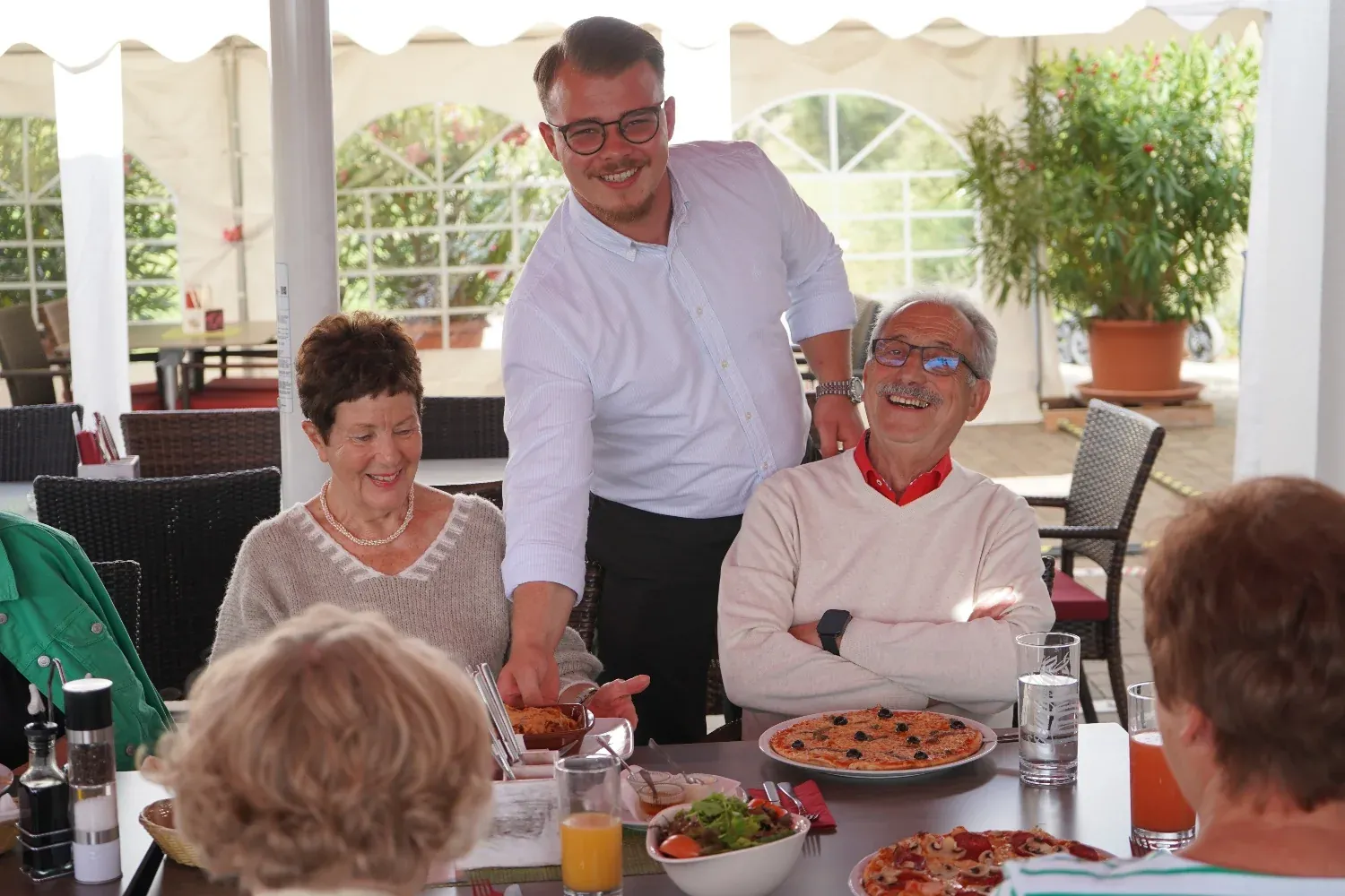 Ein Kellner serviert zwei lachenden Gästen auf der überdachten Terrasse, Pizza und Salat auf dem Tisch